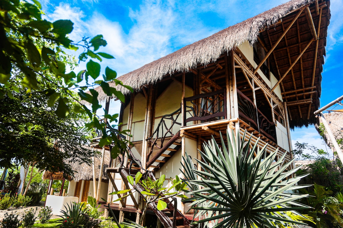 A two-story structure with a thatched roof is surrounded by lush greenery. A wooden staircase leads to the upper level, while large windows provide glimpses of the inviting interior. Unique plants and landscaping add to the charm of this tropical retreat.