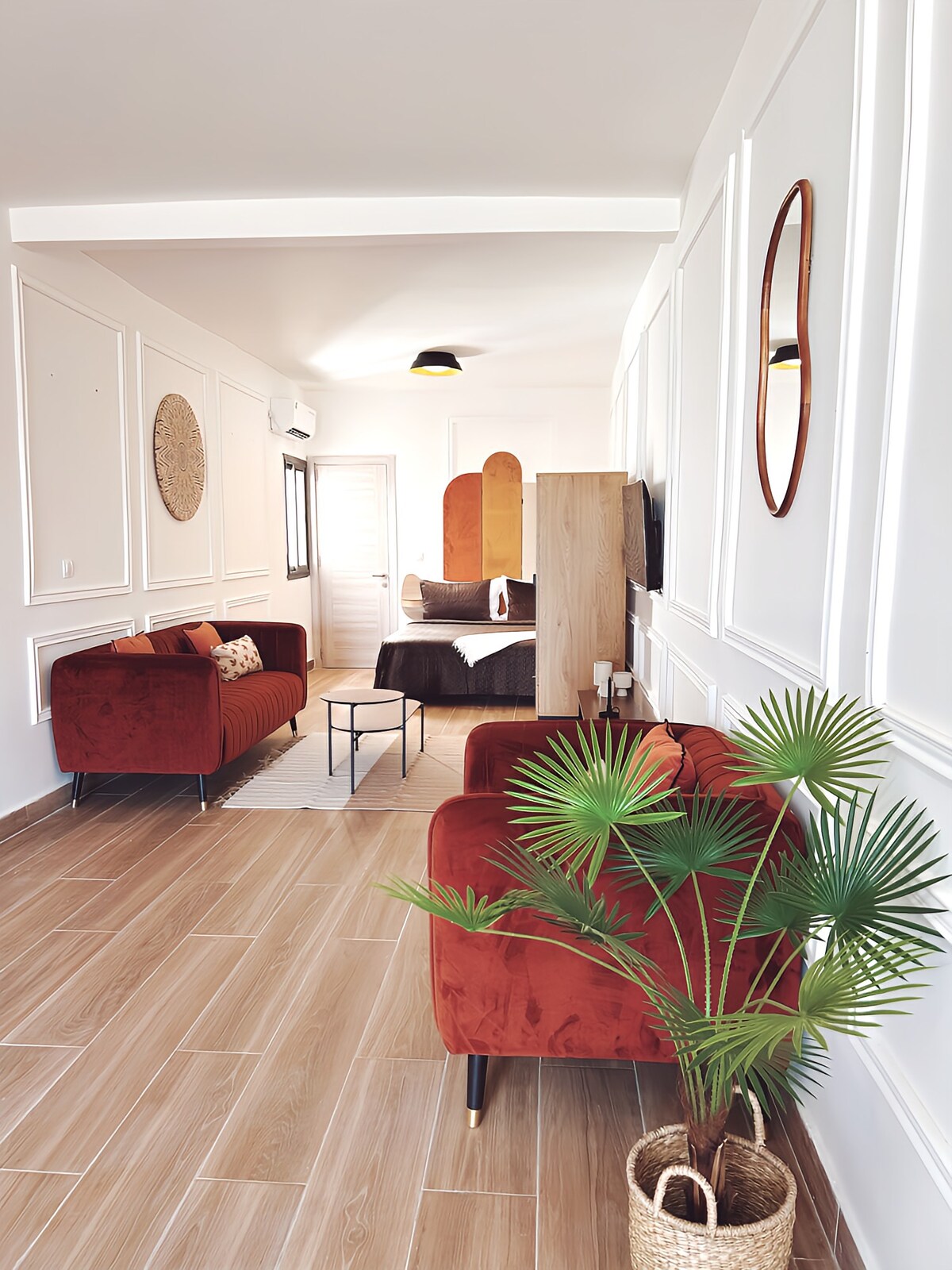 A bright living area features two red sofas facing each other, complemented by a small black table. A palm plant in a woven basket adds a touch of greenery. The space is connected to a bedroom area, showcasing light-colored walls and a modern decor style.