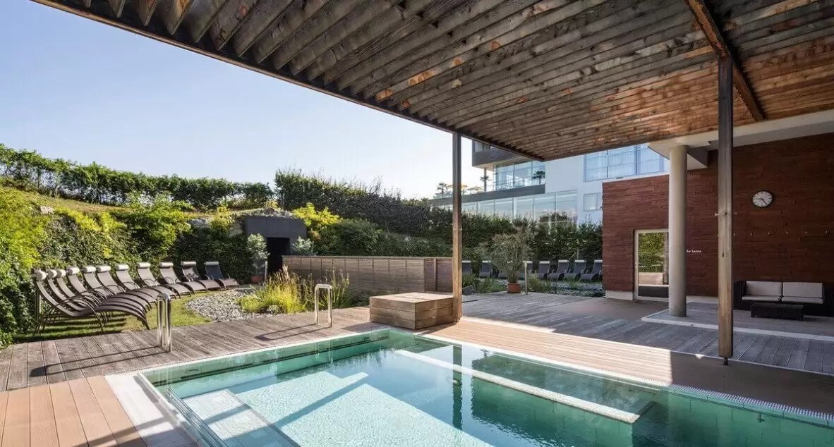 An outdoor wellness area features a rectangular pool surrounded by wood decking. Lounge chairs are positioned neatly under a shaded pergola, with lush greenery in the background. A clock is mounted on a nearby wall, and the space is brightened by ample natural light.