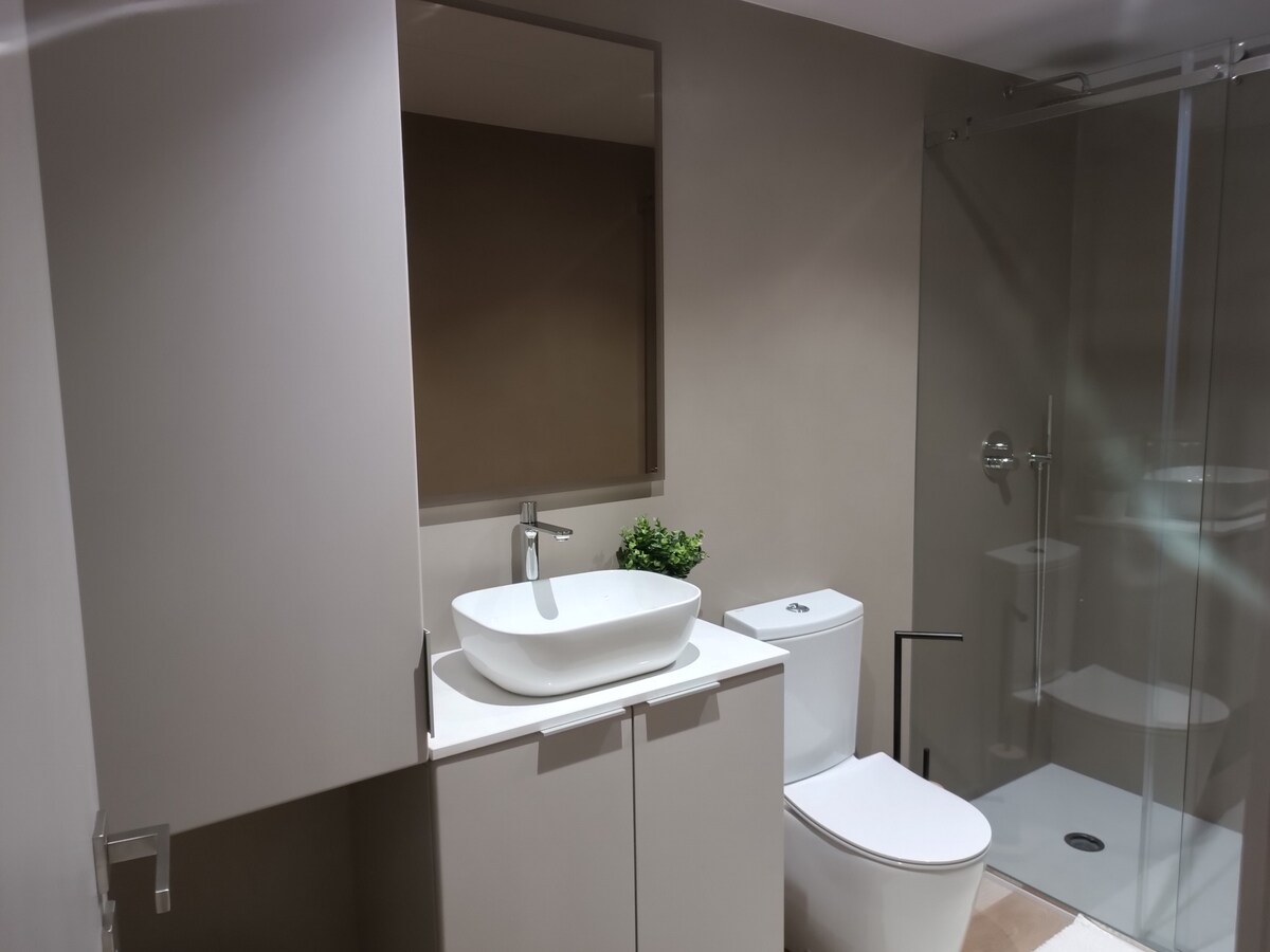 A modern bathroom is displayed, featuring a sleek countertop basin above a minimalistic vanity. A glass-enclosed shower area is visible, complemented by a toilet positioned nearby. Neutral tones dominate the space, creating a clean and functional atmosphere.