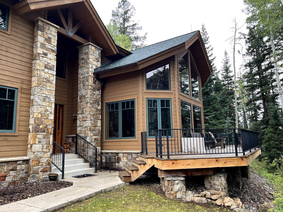 The exterior of a rustic home is depicted, featuring a blend of stone and wood elements. Large windows provide ample natural light, showcasing a spacious deck with seating. Surrounding trees enhance the natural setting, creating a serene atmosphere in a mountainous environment.