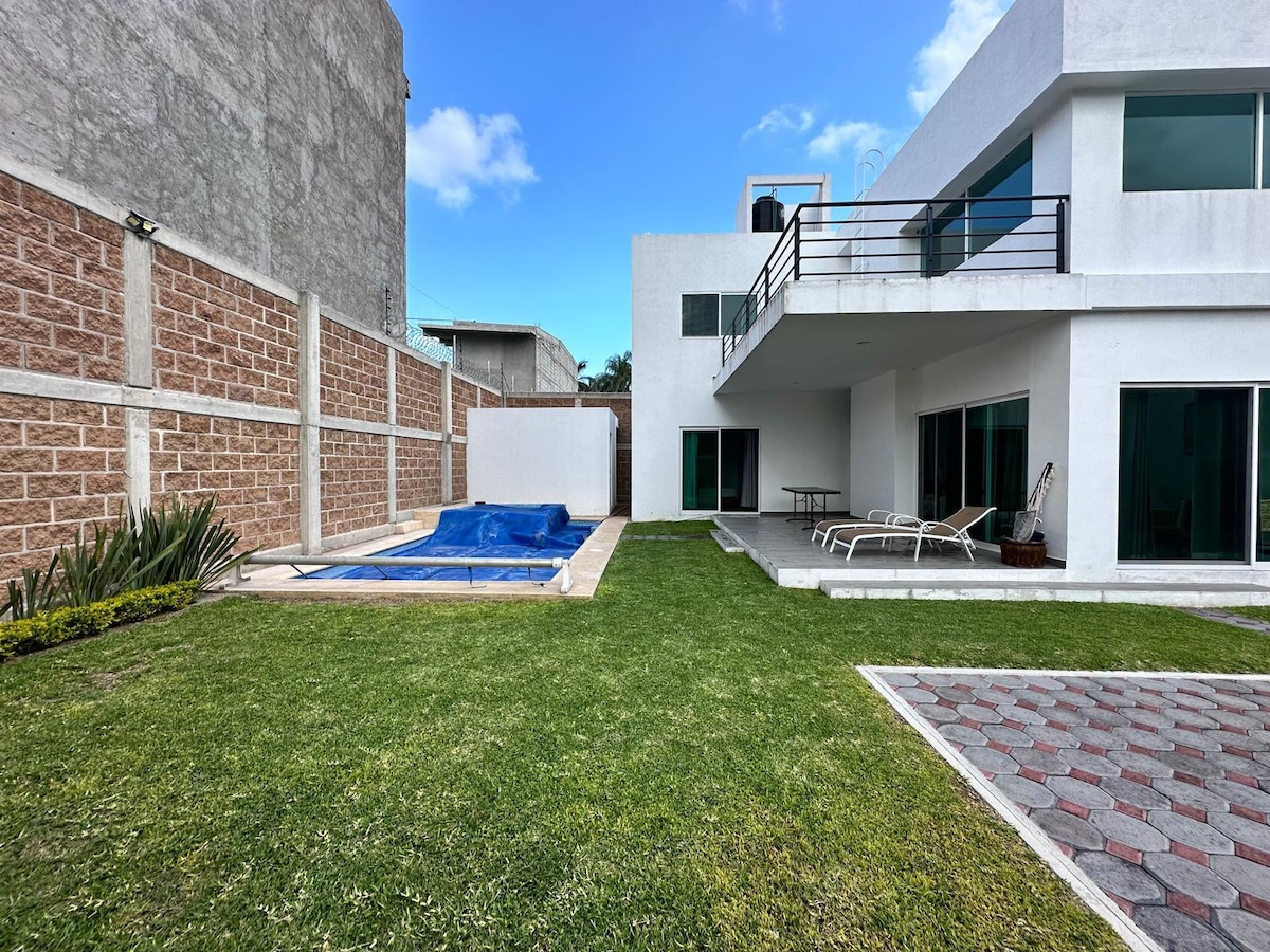 An outdoor space is presented with a private pool surrounded by grass. A terrace is visible on the second floor, offering views of the yard. Lounge chairs are arranged along the poolside. The facade features large glass doors that provide access to the interior.
