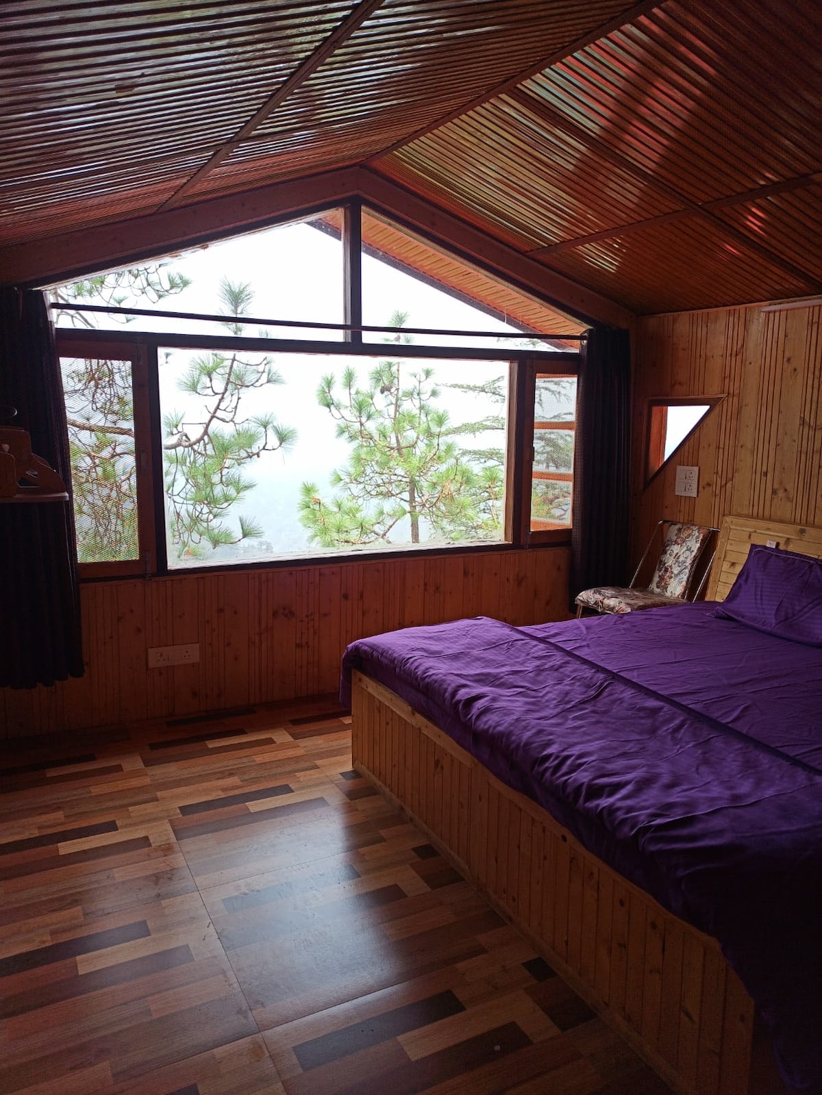 A cozy bedroom features a spacious bed covered with purple bedding. Large windows provide a view of the surrounding pine trees and mist, while wooden paneling enhances the warm atmosphere. A comfortable chair adds an additional seating option near the window.