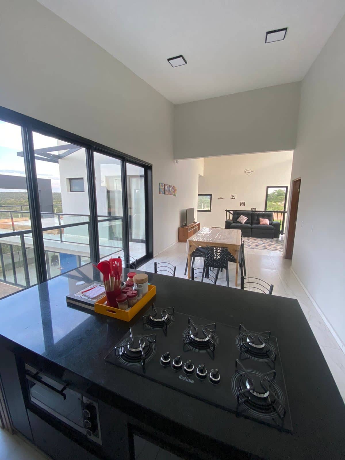 An open-concept living space is visible, featuring a modern kitchen with a dark countertop and a gas stove. A dining area with a table and chairs is set in the foreground, while large glass doors lead to an outdoor view, bringing natural light into the room.