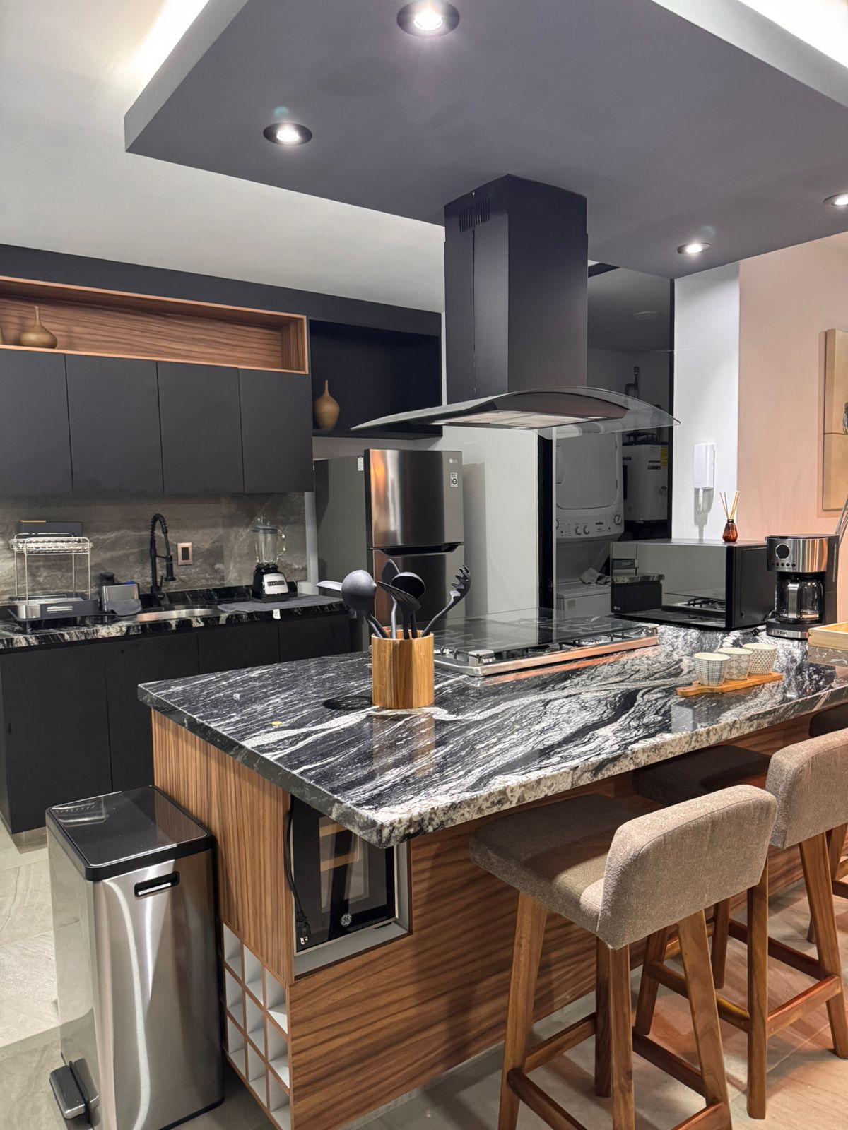 A modern kitchen features a sleek granite countertop with bar seating for three. High-end appliances, including a refrigerator and stove, are integrated into the cabinetry. Soft lighting illuminates the space, highlighting organized cookware and utensils.