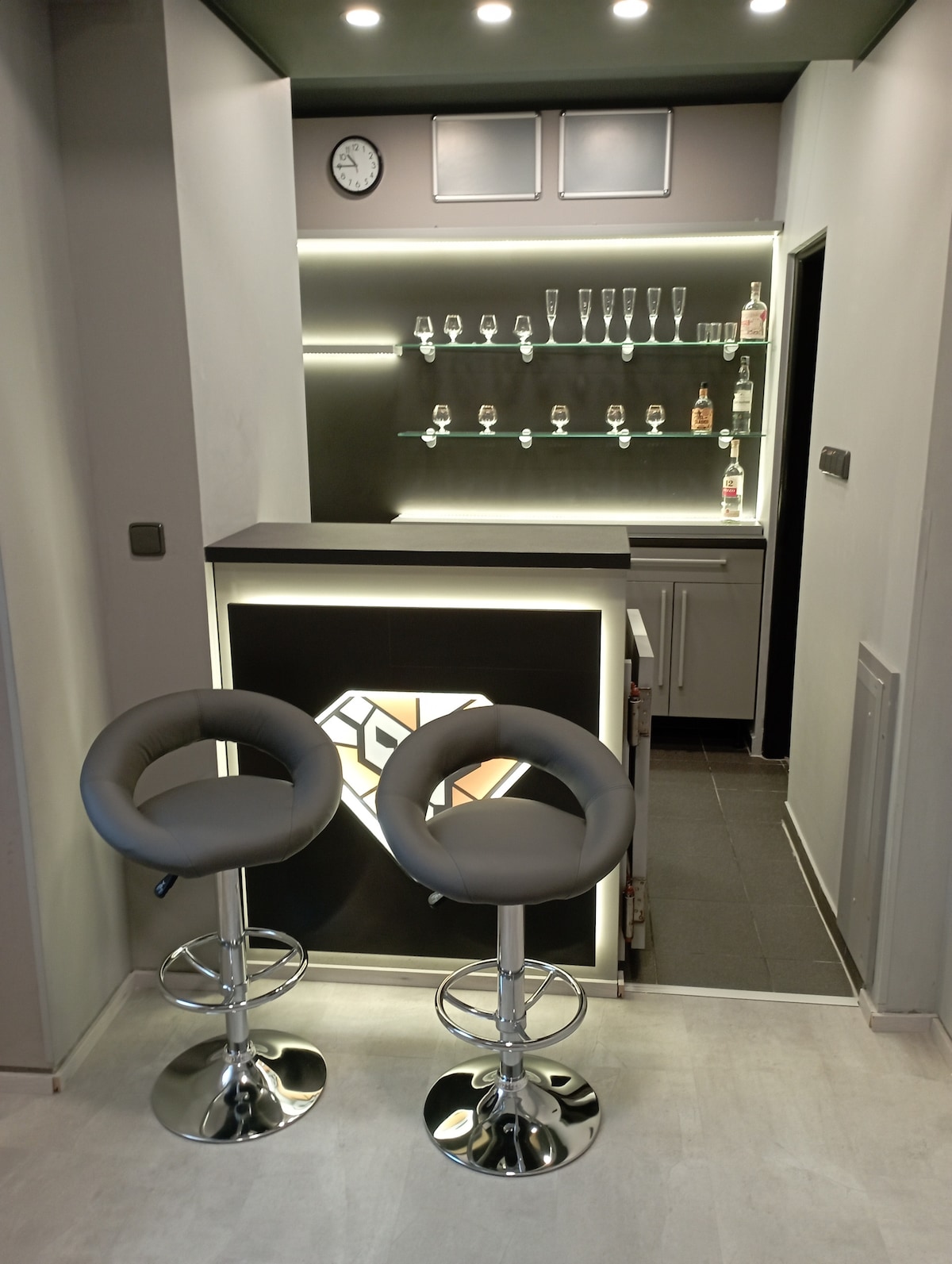 A modern bar area features a sleek countertop and a well-organized display of glasses on a shelf. Two adjustable bar stools with circular seats stand in front of the bar, and a clock is mounted on the wall. Subtle lighting highlights the space.
