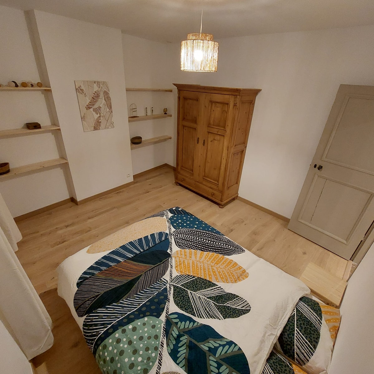 A cozy bedroom features a spacious bed with a vibrant quilt displaying a leaf pattern. An antique wooden wardrobe adds character, while open shelving holds decorative items. Soft lighting from a pendant fixture complements the warm wooden flooring and neutral wall colors.