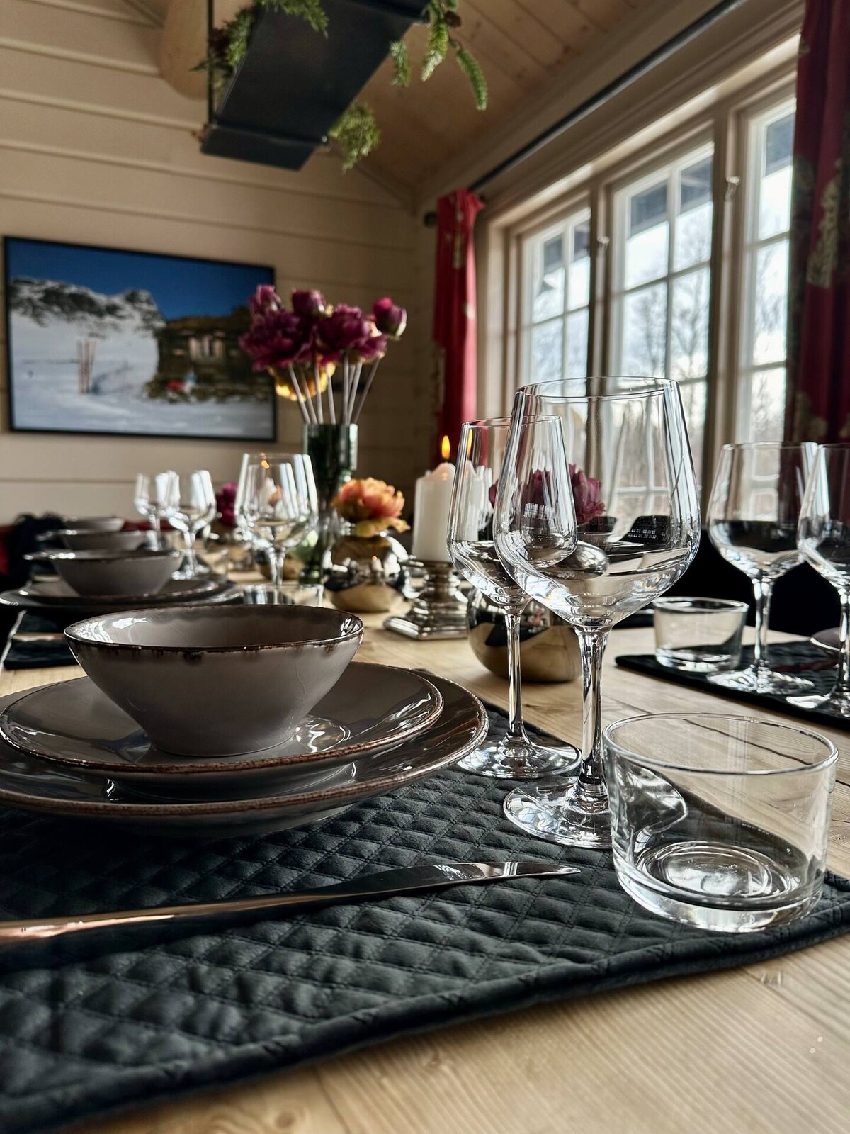 A beautifully set dining table features an array of glassware, including wine glasses and smaller cups, arranged beside elegant plates. Soft lighting enhances the cozy ambiance, while flowers are displayed nearby. Large windows invite natural light and showcase a scenic outdoor view.