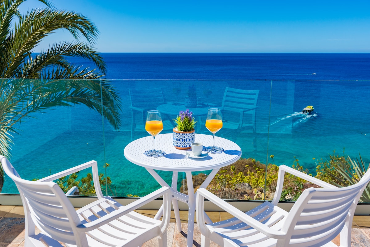 A glass railing encloses a small terrace, offering uninterrupted views of the ocean. Two white chairs and a round table are positioned to enjoy the scenery, adorned with a potted plant and two glasses of orange beverage, reflecting bright sunlight.
