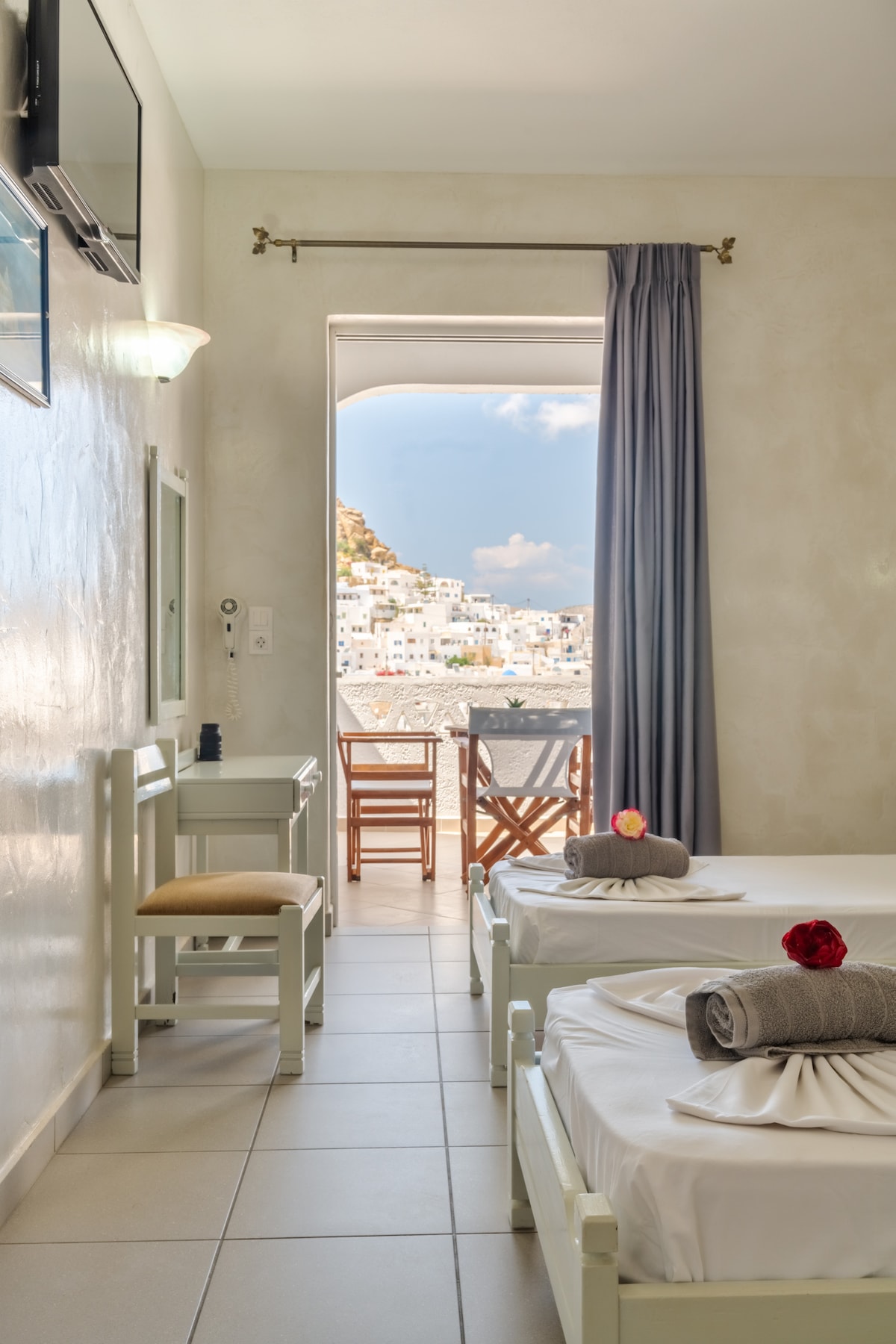 A room features two spacious beds, each adorned with towels and decorative flowers. A small work desk and chair are positioned near a window, which opens to a private balcony offering a view of the surrounding landscape. Soft natural light fills the space.