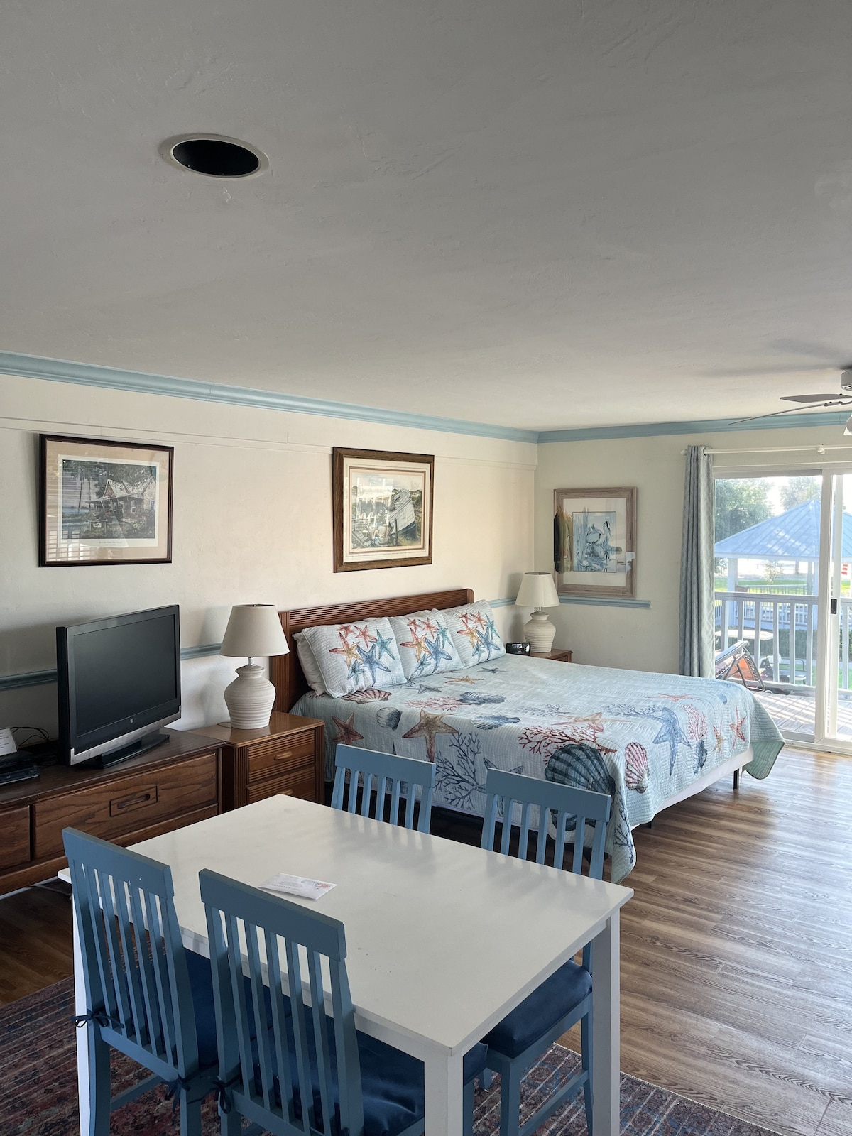 A compact living space features a queen-sized bed adorned with a nautical-themed quilt. A wooden dining table with four chairs occupies the area. A television is placed on a low cabinet. Natural light fills the room through sliding glass doors leading to a balcony.