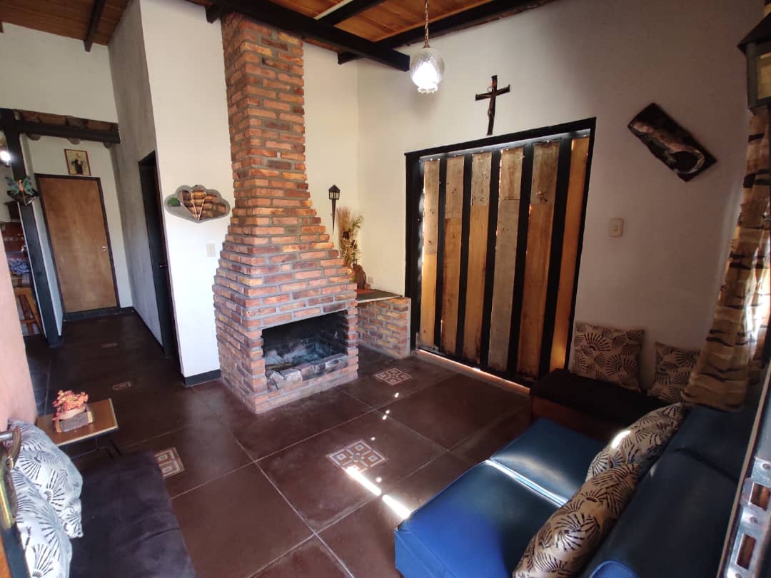 A cozy living area features a rustic brick fireplace as a centerpiece. Exposed wooden beams create a warm ambiance. Two comfortable sofas are positioned to invite relaxation, while a wooden cross and decorative elements add character to the space. Natural light filters through the nearby door.