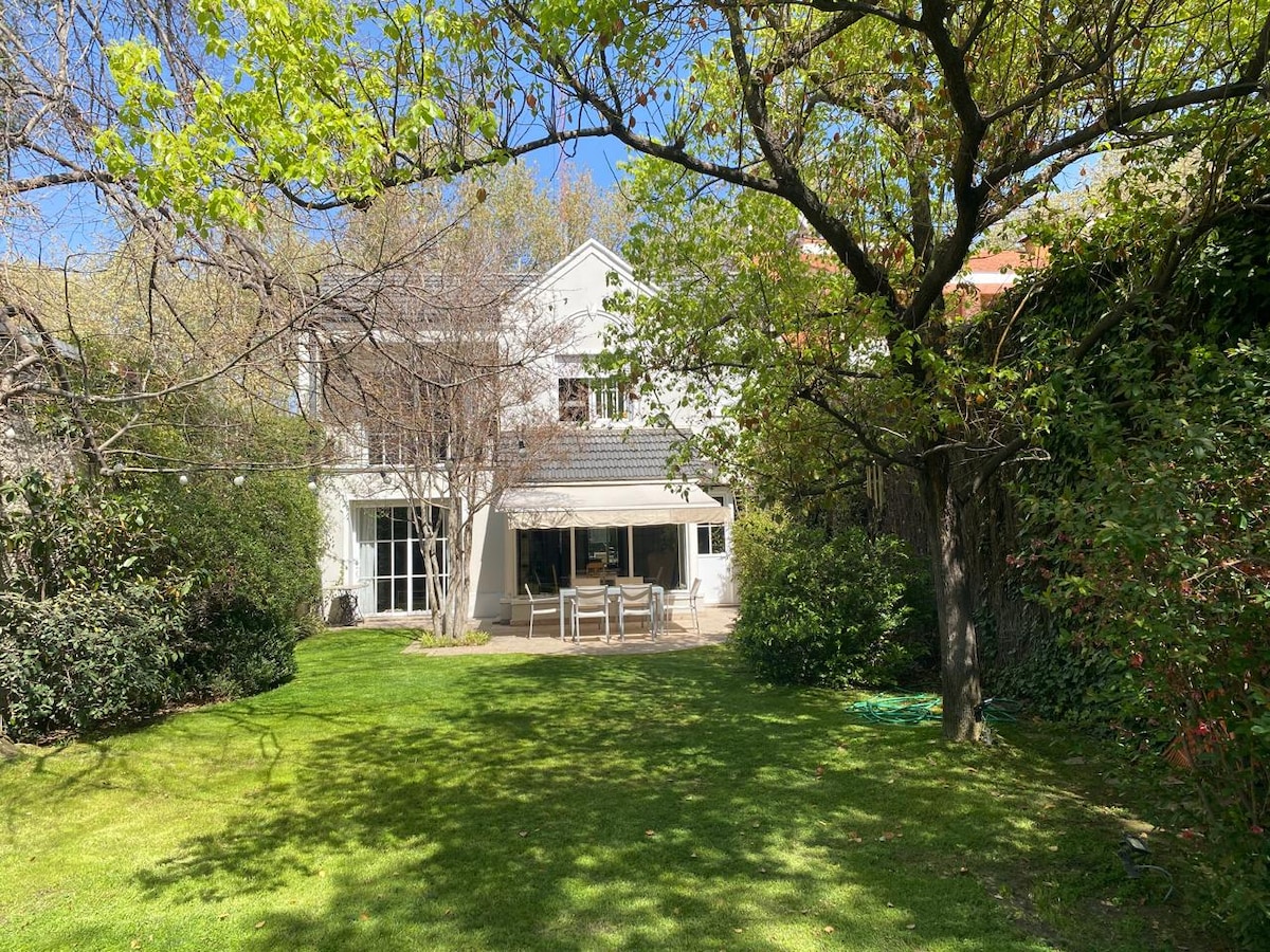 A spacious backyard features a well-maintained lawn surrounded by trees, providing shade and greenery. A patio area is visible, furnished with dining chairs and a table, and a series of large windows allow views of the interior from the outside.
