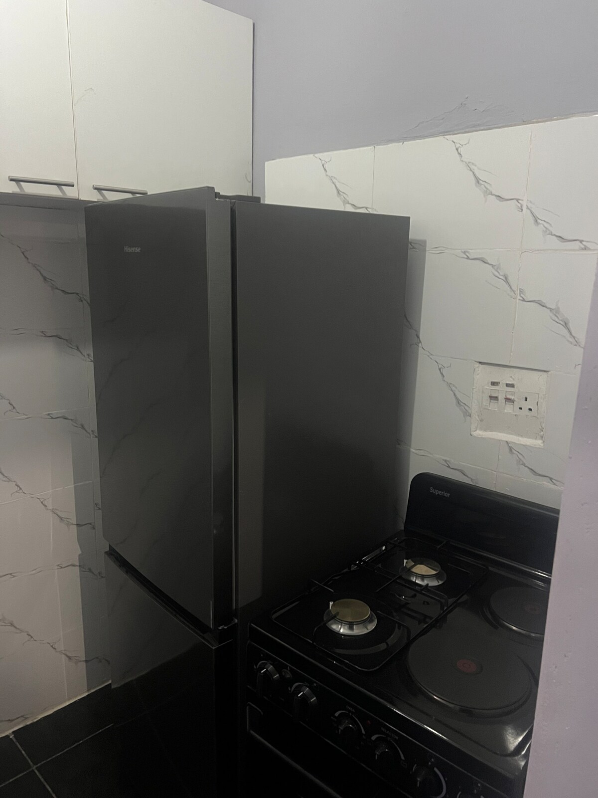 A compact kitchen area features a sleek black refrigerator adjacent to a gas stove. The appliances are set against a backdrop of light-colored tiled walls with a marble pattern, providing a modern touch to the space.