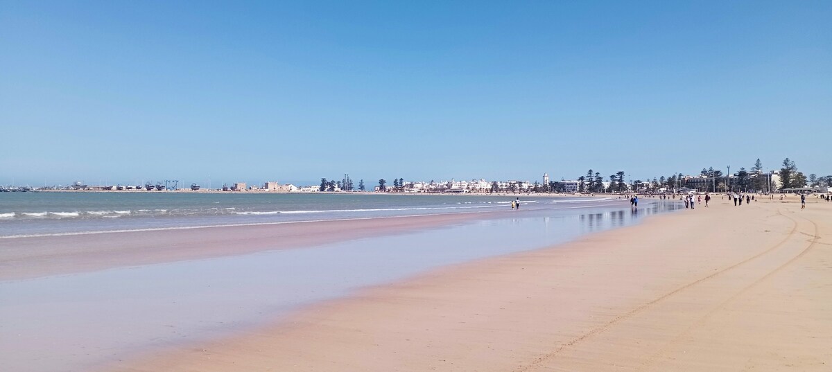A wide sandy beach stretches along the coastline, with the gentle waves lapping at the shore. In the distance, the town's buildings are visible, framed by palm trees under a clear blue sky. People can be seen enjoying the beach along the water's edge.