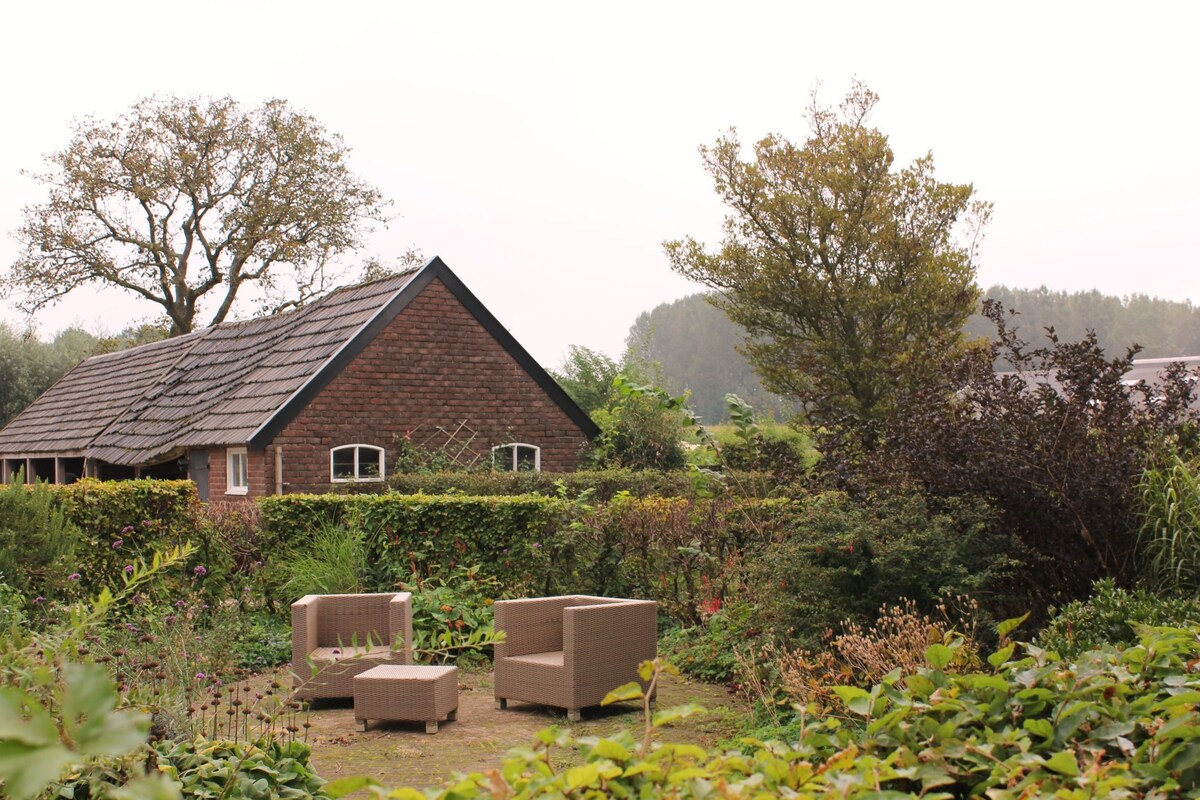 A cozy outdoor seating area is nestled within a lush garden, featuring two woven chairs and a small table. Surrounding greenery provides a sense of privacy, while a charming farmhouse with a rustic roof is visible in the background, complementing the serene atmosphere.