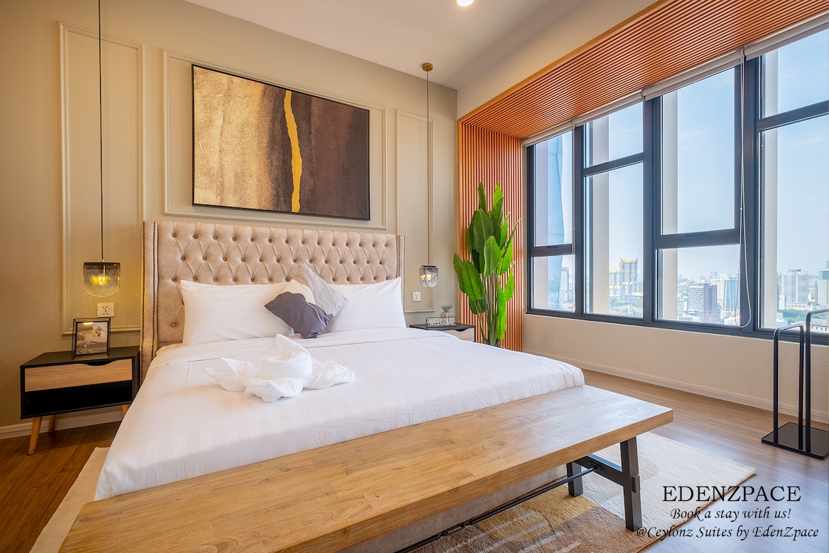 A spacious bedroom features a comfortable bed with neutral linens and a headboard designed in a tufted style. Large windows provide ample natural light, offering a view of the city skyline. A decorative plant and a bedside table are present, creating a serene environment.