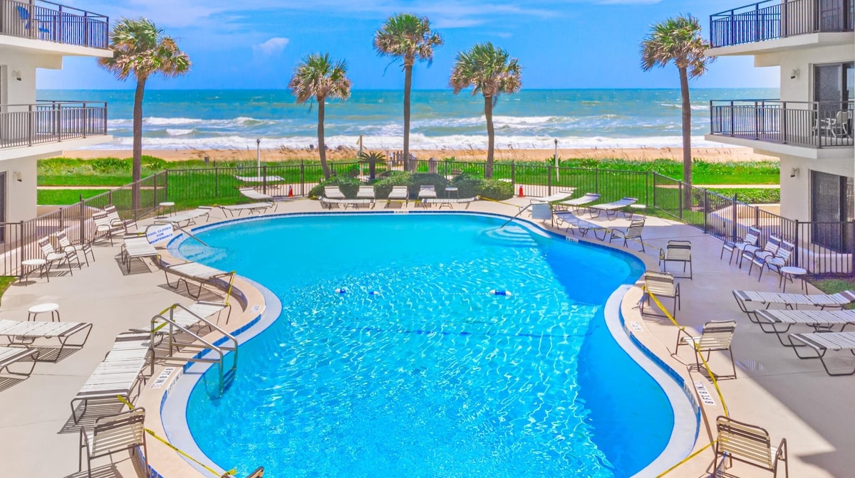 A large, inviting swimming pool occupies the foreground, surrounded by lounge chairs. Palm trees sway gently nearby, framing the view of the ocean beyond. The tranquil water reflects the sky, enhancing the appeal of the beachfront setting.
