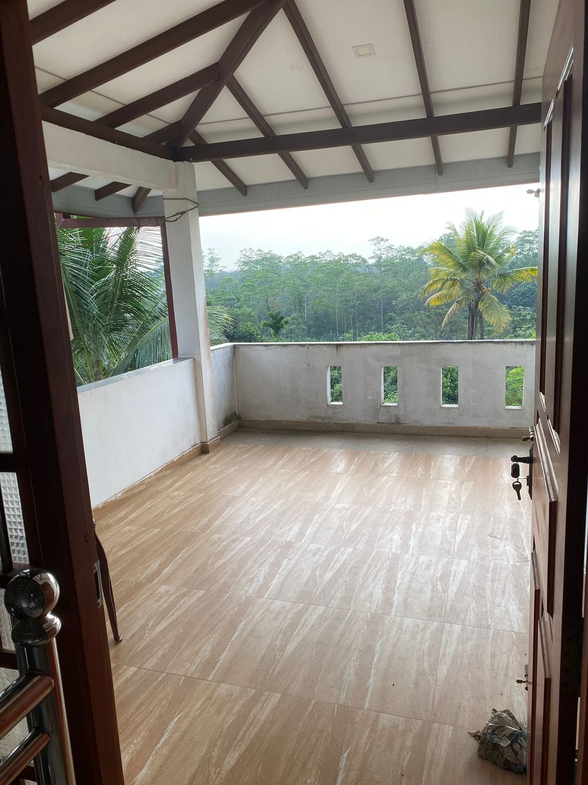 A spacious balcony extends from the entrance, featuring tiled flooring and framed by a wooden railing. Lush greenery is visible in the background, with tall trees and plants enhancing the serene setting. Natural light floods the area, creating an inviting and airy feel.
