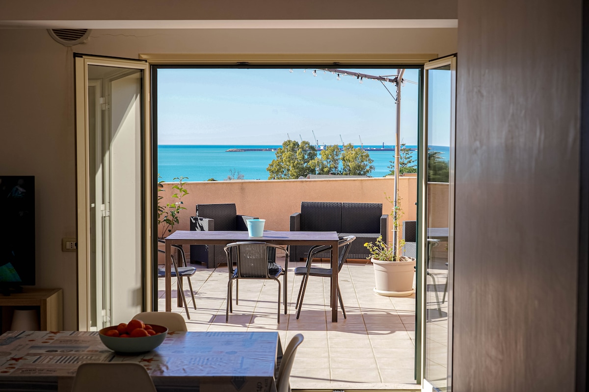 An open doorway reveals a terrace with outdoor seating, overlooking a clear blue sea. Sunlight brightens the interior space, which includes a dining table and chairs. Potted plants are positioned on the terrace, enhancing the coastal view.