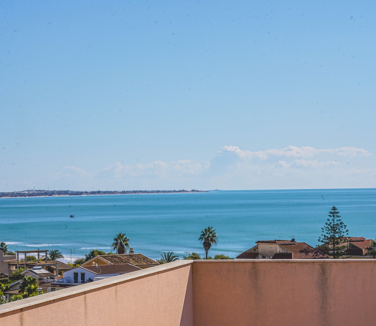 A panoramic view of the Mediterranean Sea is captured from the terrace, showcasing clear blue waters and a distant horizon. Palm trees and rooftops of nearby buildings are visible, creating a serene coastal atmosphere.