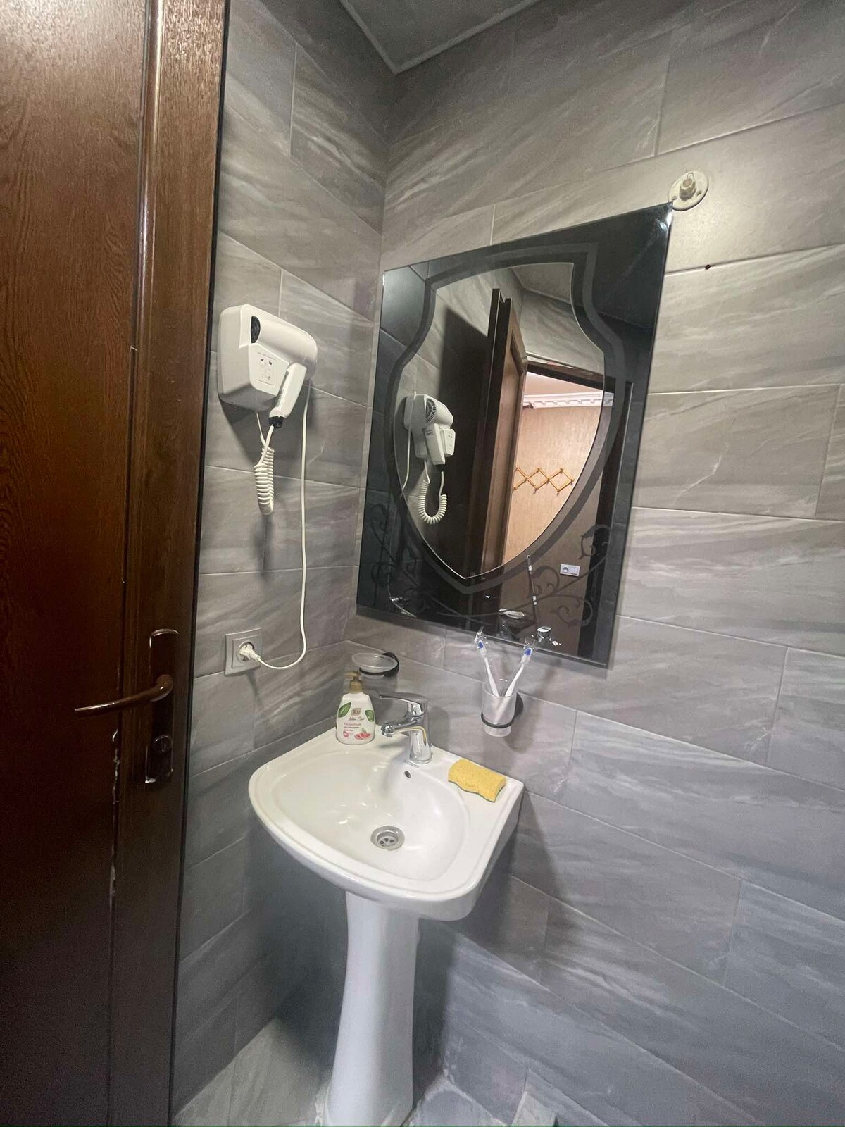 A compact bathroom is presented, featuring a sleek sink mounted on a pedestal. Gray tiles line the walls, contributing to a modern look. Above the sink, a dark-framed mirror provides reflection, while a hairdryer is conveniently placed nearby, enhancing functionality.