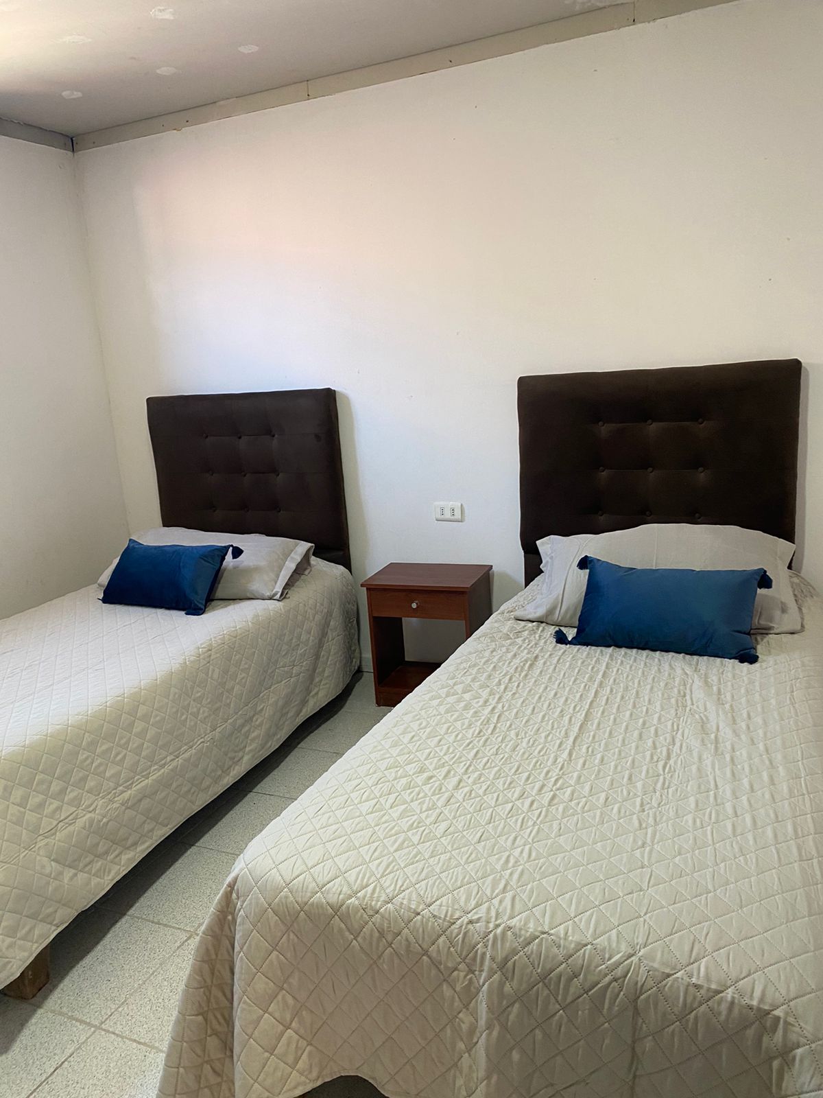 A simple and clean bedroom features two spacious beds with tufted headboards. Each bed is dressed in neutral quilts and paired with blue decorative pillows. A small bedside table is positioned between the beds, enhancing the functional layout.