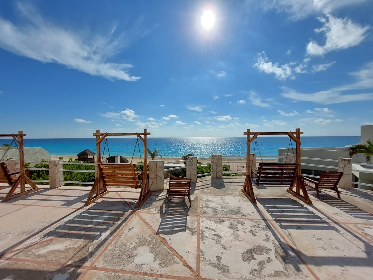 A spacious terrace is presented with wooden swings facing the sea, allowing for relaxation and scenic views. The clear blue sky is filled with soft clouds, and sunlight glistens on the water, creating a serene atmosphere. The terrace floor features a light-colored tiling pattern.