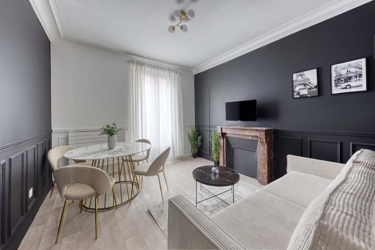 A modern living area features a neutral color palette with dark accent walls. A round dining table is surrounded by four light-colored chairs. A marble fireplace serves as a focal point, complemented by two framed black-and-white photographs. Natural light enters through the window, enhancing the space.