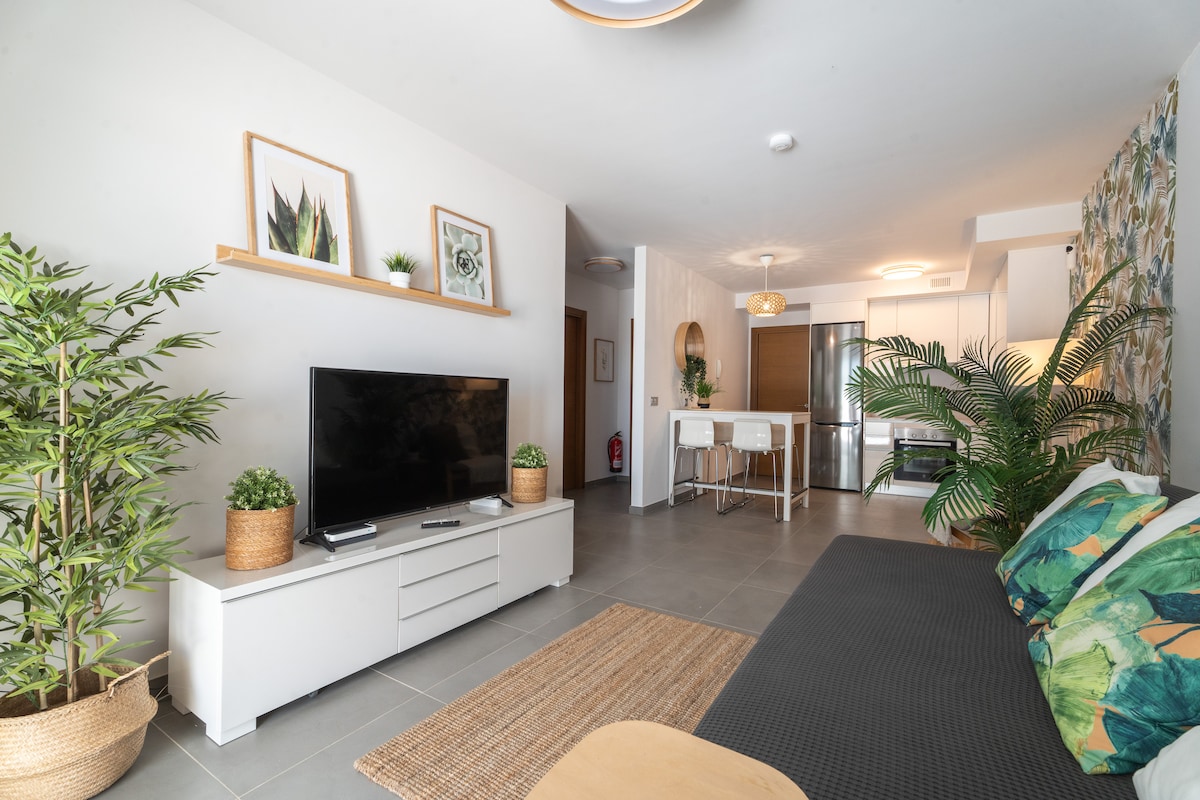 A living area is presented with a flat-screen TV on a white cabinet. Green plants are positioned on either side of the TV. A dark sofa features decorative cushions, and a jute rug is underfoot. A dining area is visible in the background, furnished with white chairs and a round table.