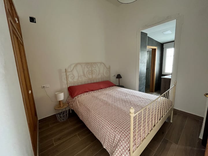 Dormitorio acogedor con cama de forja blanca y colcha geométrica. Cuenta un amplio armario empotrado, el suelo cerámico imitación madera y acceso directo a un baño moderno con azulejos oscuros, creando una suite luminosa y funcional.