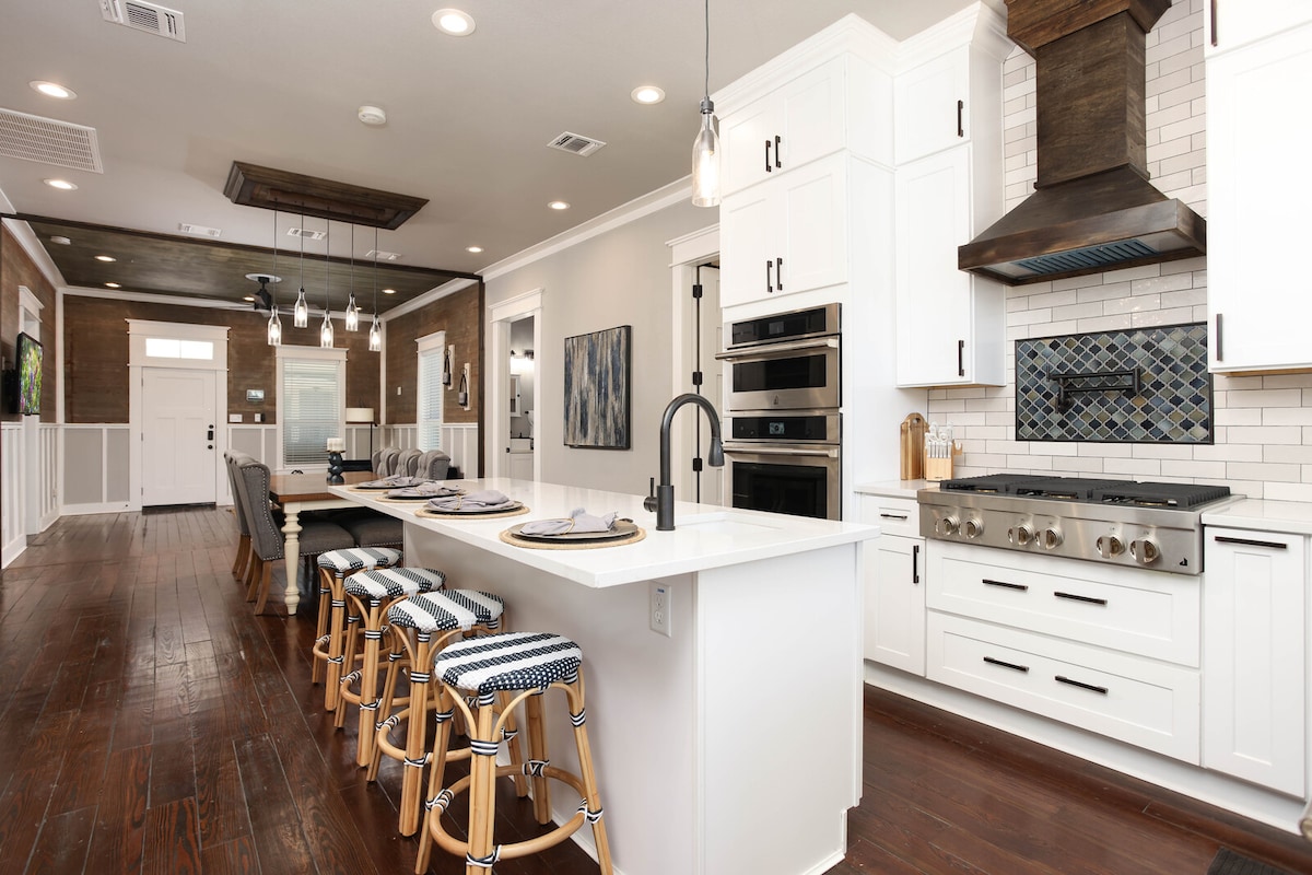 A spacious chef's kitchen is presented, featuring modern Jenn-Air appliances, including a six-burner gas stove and a large island with seating for four. White cabinetry and a striking backsplash contribute to the bright atmosphere, while hardwood floors add warmth to the space.