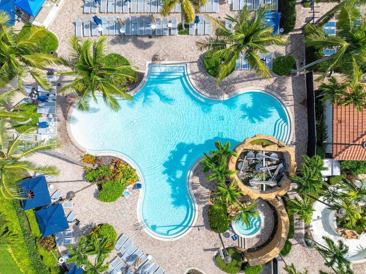 Aerial view of a uniquely shaped swimming pool surrounded by palm trees and lounging chairs. A decorative waterfall feature is visible at one end, adding a touch of elegance to the serene environment. Sun umbrellas provide shaded areas around the pool.