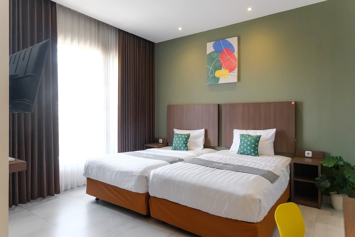A well-lit room features two spacious beds with white linens and green accent pillows. A colorful abstract wall art is displayed above the beds. Natural light filters through sheer curtains, complementing the green accent wall and creating a soothing environment.