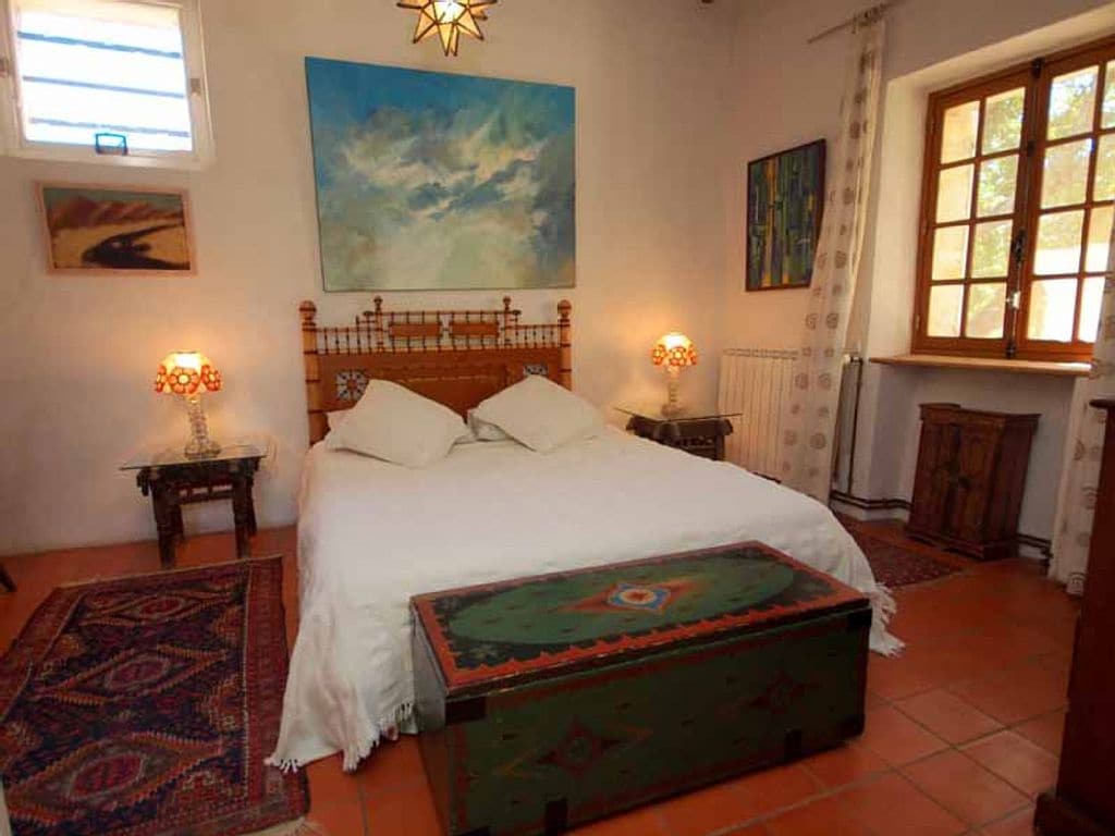 The spacious room features a double bed with crisp white linens, complemented by decorative pillows. Two bedside lamps illuminate the space, while a wooden chest with intricate designs lies at the foot of the bed. Natural light enters through a window adorned with sheer curtains.