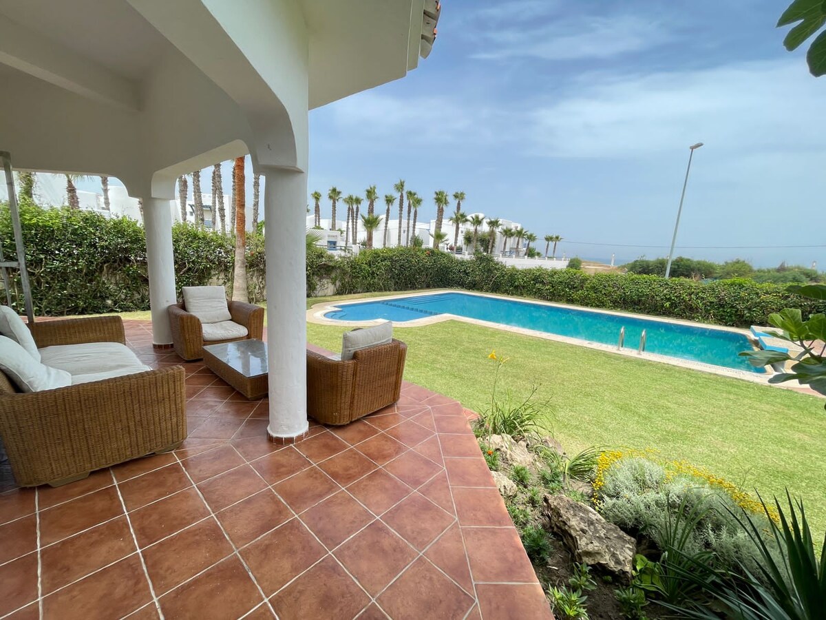 An outdoor seating area is showcased with wicker chairs and a glass table, bordered by a well-maintained lawn. A serene swimming pool glistens in the background, surrounded by palm trees under a clear blue sky.