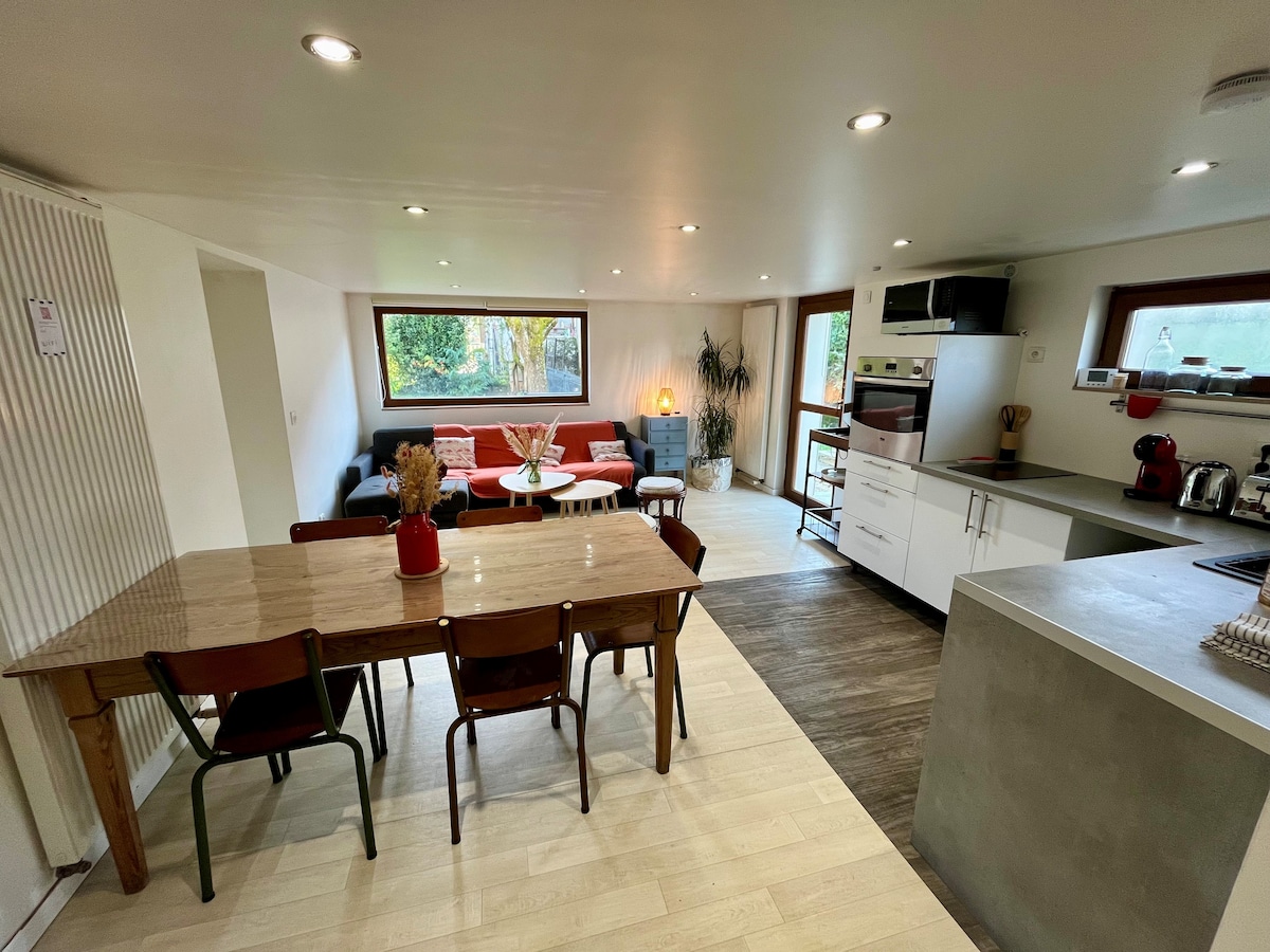 An open living space features a dining table surrounded by four chairs, a cozy seating area with a red sofa, and a minimalist kitchen with modern appliances. Large windows allow natural light to brighten the interior, while decorative plants add a touch of greenery.