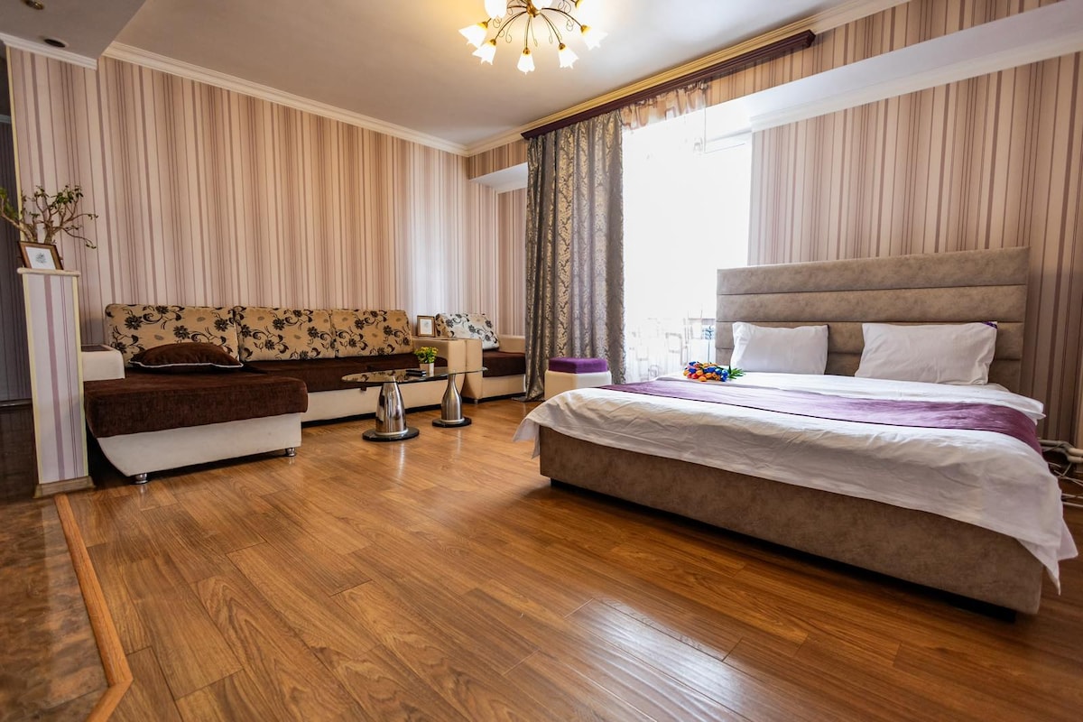 The spacious bedroom features a comfortable bed dressed in neutral tones. A seating area with patterned upholstery is present, complemented by a coffee table. Natural light filters through the window, highlighting the warm wooden flooring and striped wallpaper.