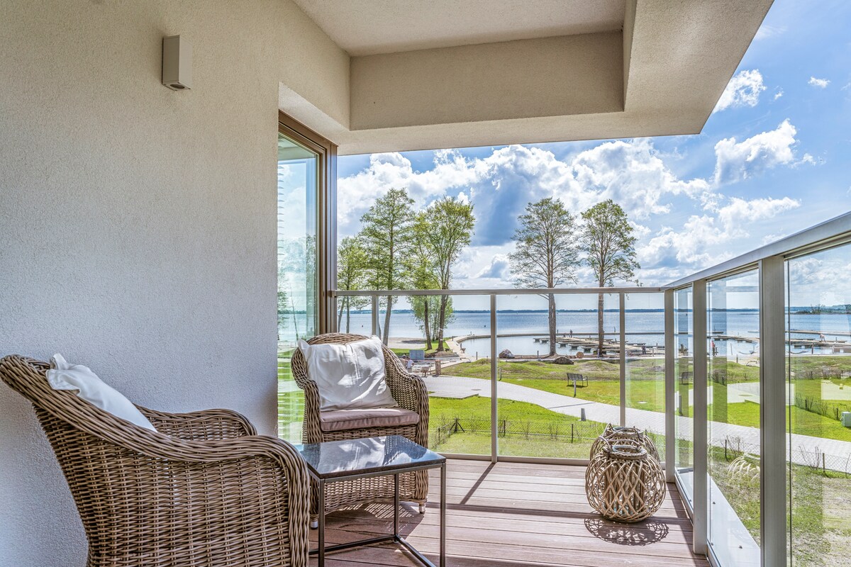 A private balcony is shown, featuring two woven chairs and a glass table. The view presents lush greenery and a glimpse of the lake under a partly cloudy sky, enhancing the outdoor experience.