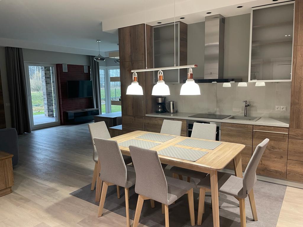 A modern kitchen and dining area are displayed, featuring a wooden dining table with six upholstered chairs. Above, three pendant lights provide illumination. The kitchen is equipped with sleek cabinetry and contemporary appliances, while large windows allow natural light and views of the garden.