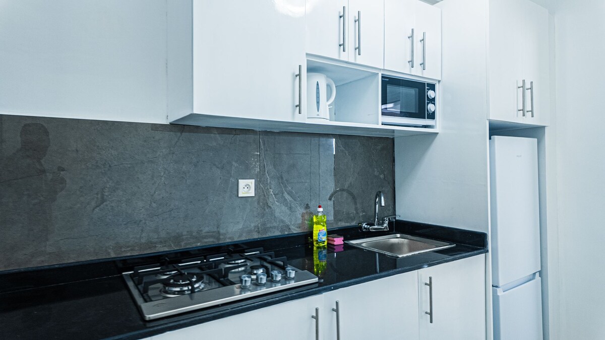 The kitchen features modern cabinetry in a sleek white finish, enhanced by a dark stone backsplash. A gas stove, microwave, and refrigerator are included, alongside a stainless steel sink. A bottle of dish soap and a mug are placed on the countertop.