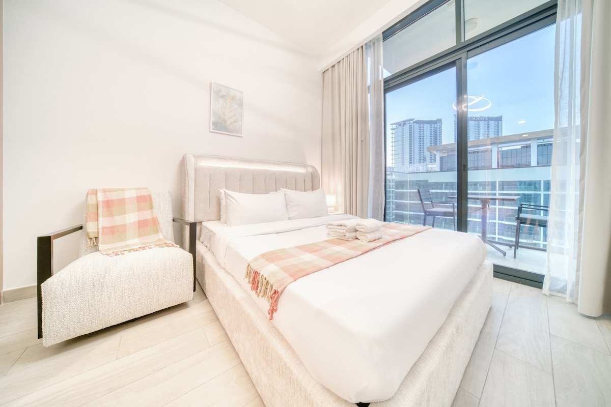 A well-lit bedroom features a spacious bed adorned with a soft throw blanket. A comfortable chair is positioned nearby, and large glass doors open to a private balcony with city views. Light curtains frame the window, allowing natural light to filter in.
