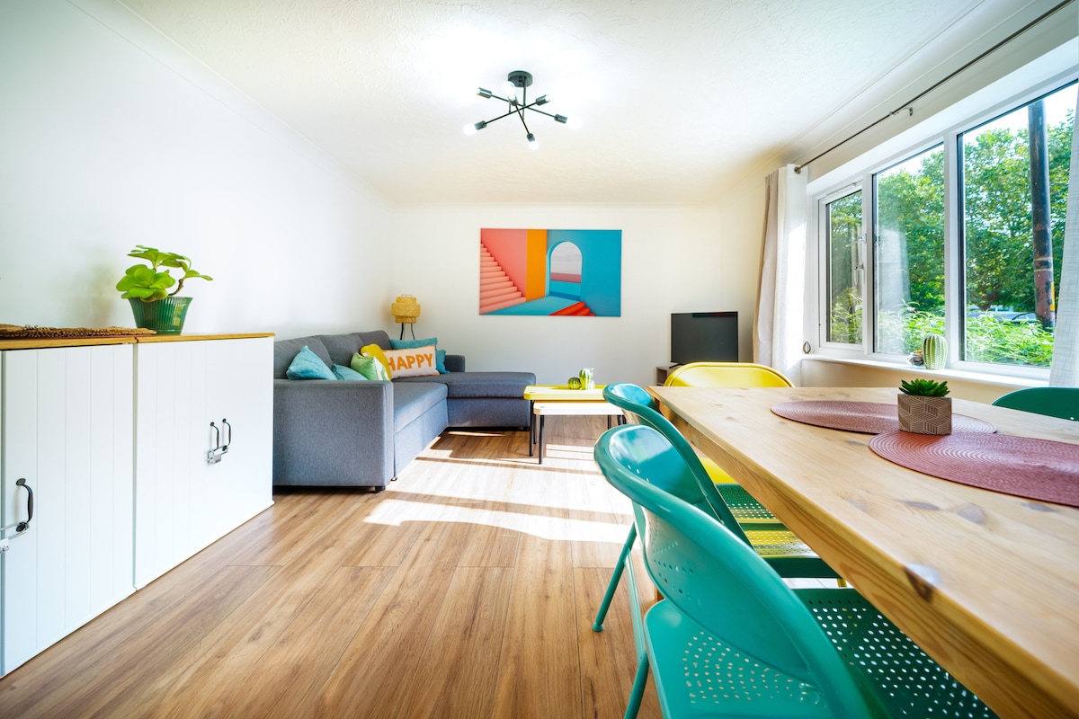 A well-lit living space features a gray sofa adjacent to a vibrant artwork. A multi-colored dining area includes a wooden table surrounded by brightly colored chairs. Large windows provide natural light and views of outdoor greenery, enhancing the welcoming atmosphere.
