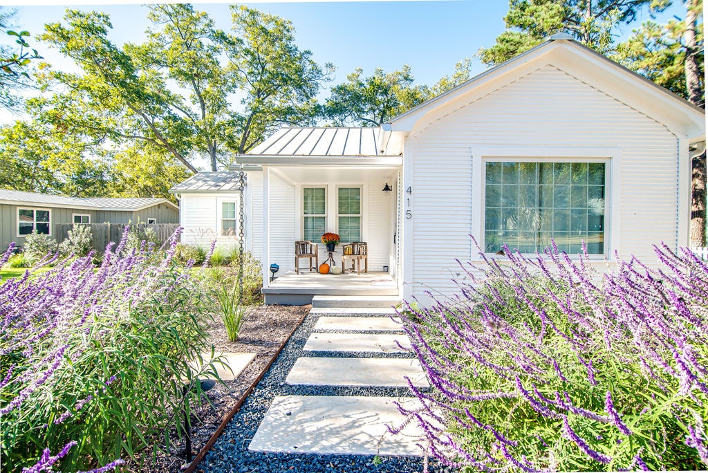 A charming cottage exterior is surrounded by vibrant purple flowers and green shrubs. A pathway leads to the inviting front porch, which features two chairs and potted plants. Sunlight creates a warm atmosphere, emphasizing the cottage's classic white siding and a metal roof.