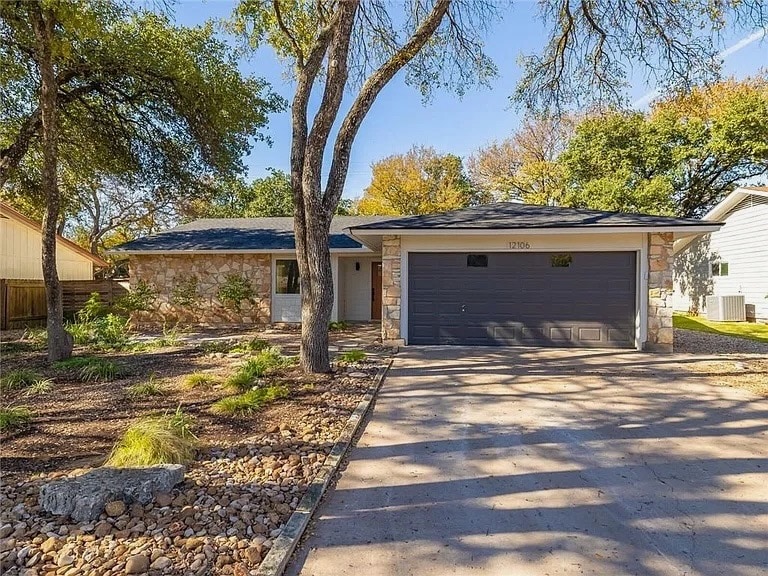 A modern single-story home is presented, featuring a stone accent exterior and a dark gray garage door. The front yard is landscaped with a rock garden and shaded by mature trees, contributing to a serene outdoor setting.