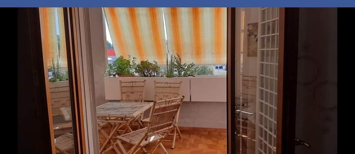 A balcony area is shown featuring a wooden table surrounded by several folding chairs. Sunlight filters through bright orange striped awnings, illuminating potted plants placed along the walls. Glass doors provide access to the space, enhancing the connection to the outdoors.
