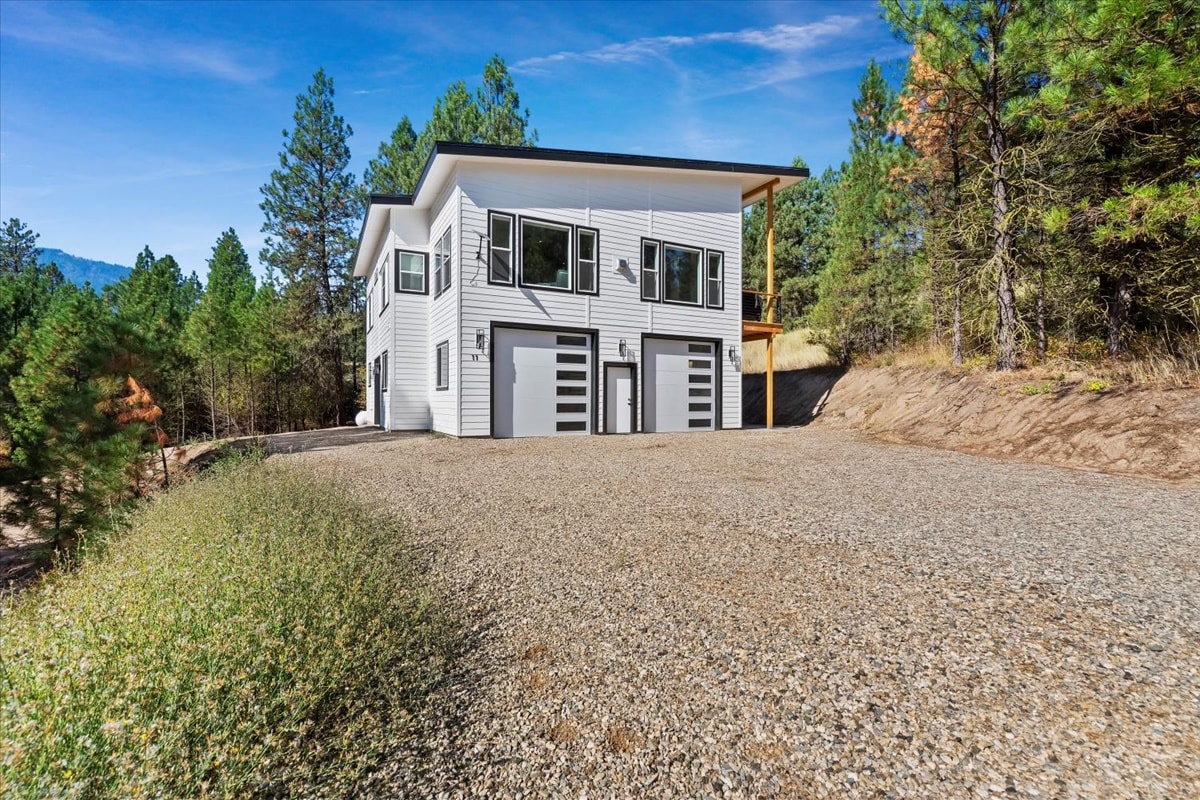 A modern two-story building is positioned amidst pine trees, featuring large windows that allow natural light to enter. The gravel driveway provides ample parking space, while the surrounding landscape emphasizes a peaceful and natural setting.