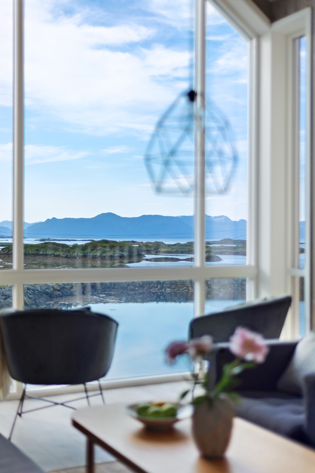 Large windows offer views of the serene landscape featuring mountains and water. A modern light fixture hangs above, while soft seating and a wooden table with fresh flowers create a welcoming indoor space that invites relaxation.