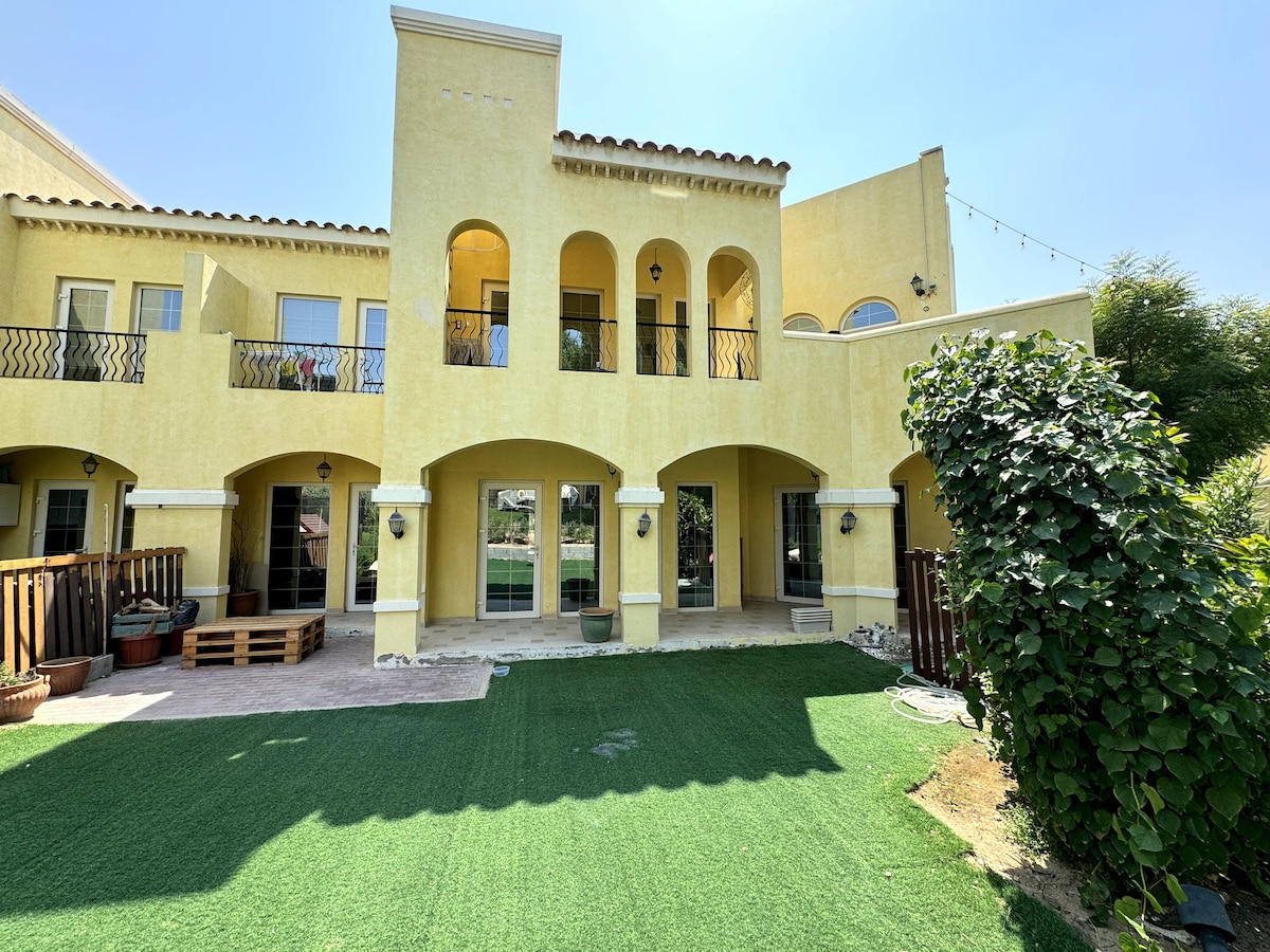 The exterior of a yellow villa features arched windows and doors, creating an inviting appearance. A well-maintained garden area with artificial grass is present, surrounded by potted plants. The structure is illuminated by natural sunlight, enhancing the welcoming ambiance of the outdoor space.