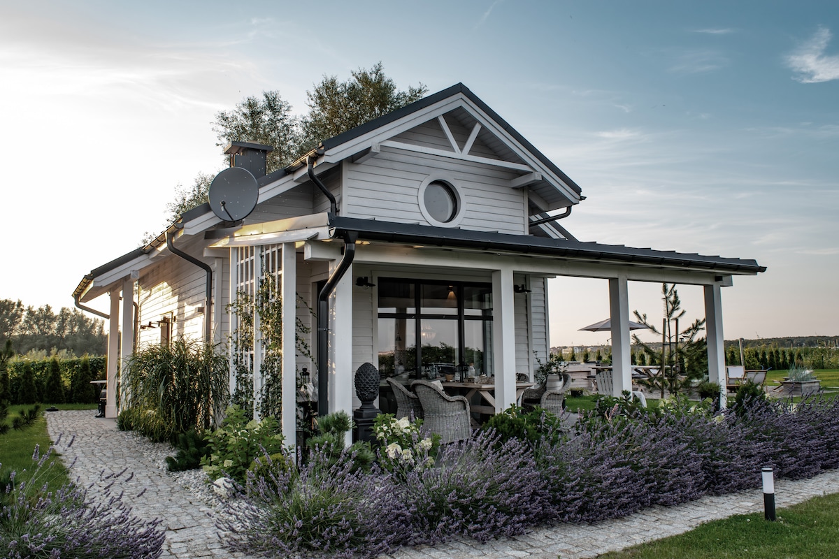 A modern single-story house is depicted, featuring a welcoming porch with comfortable seating. Surrounding greenery includes lavender plants, enhancing the outdoor space. Large windows allow ample natural light inside, while a satellite dish is mounted on the roof, indicating connectivity.